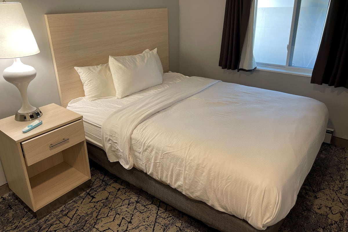 A neatly made bed with white linens, a wooden headboard, a bedside table with a lamp and remote control, and a window with curtains.