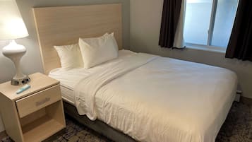 Una cama bien hecha con sábanas blancas, cabecera de madera, mesita de noche con lámpara y control remoto, y una ventana con cortinas.