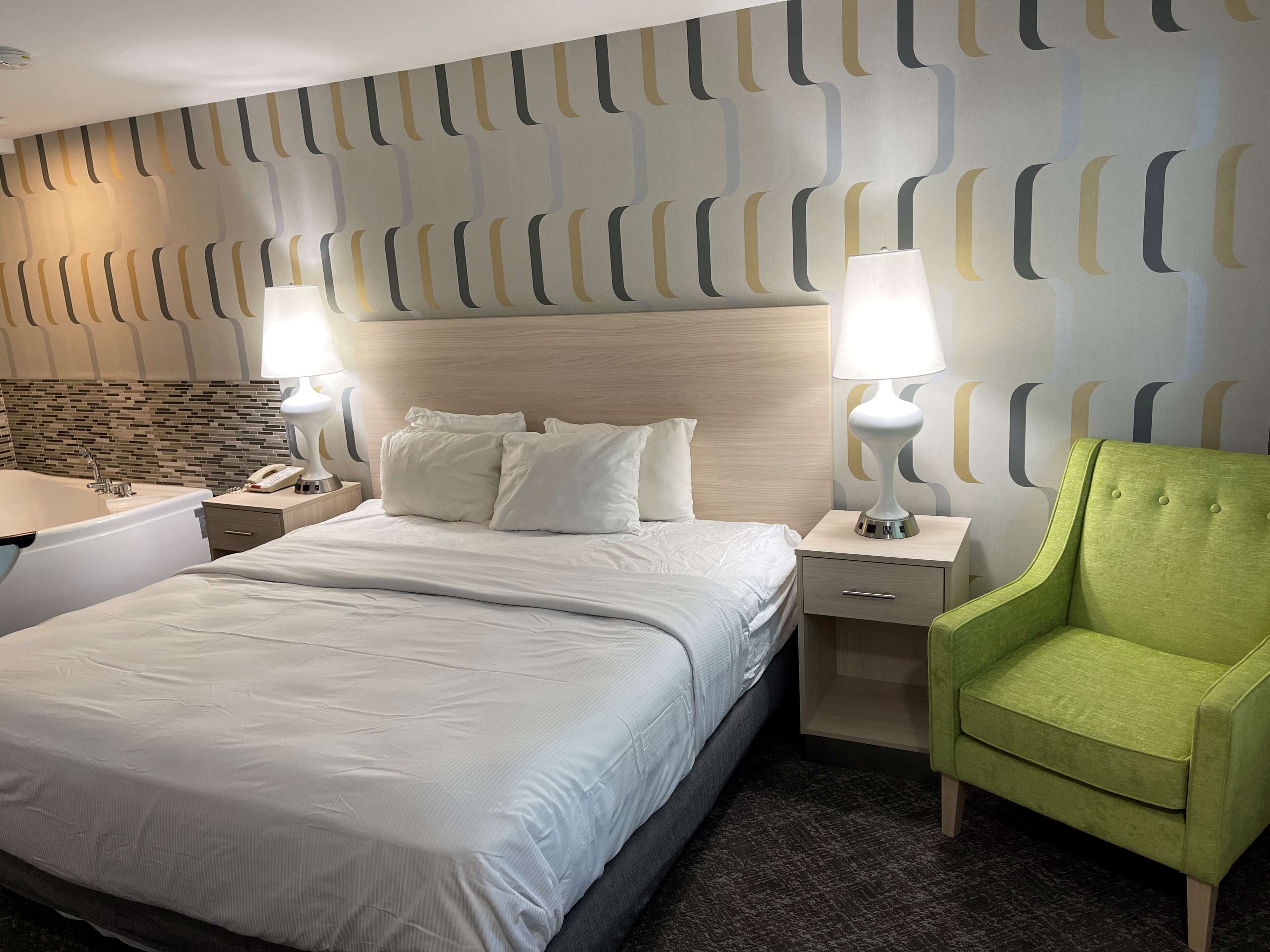 A hotel room with a large bed, two bedside lamps, a green armchair, a wooden headboard, and a patterned wall.