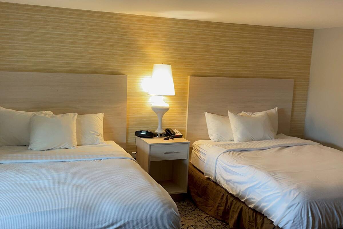A hotel room with two beds, each with white bedding and pillows. There is a nightstand between the beds with a lamp, phone, and keys.