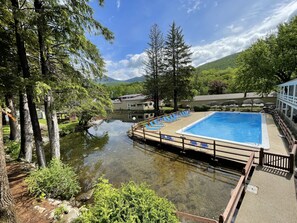 A swimming pool with a wooden deck and lounge chairs, surrounded by trees and a small body of water. - Woodwards White Mountain Resort, BW Signature Collection (Lincoln)
