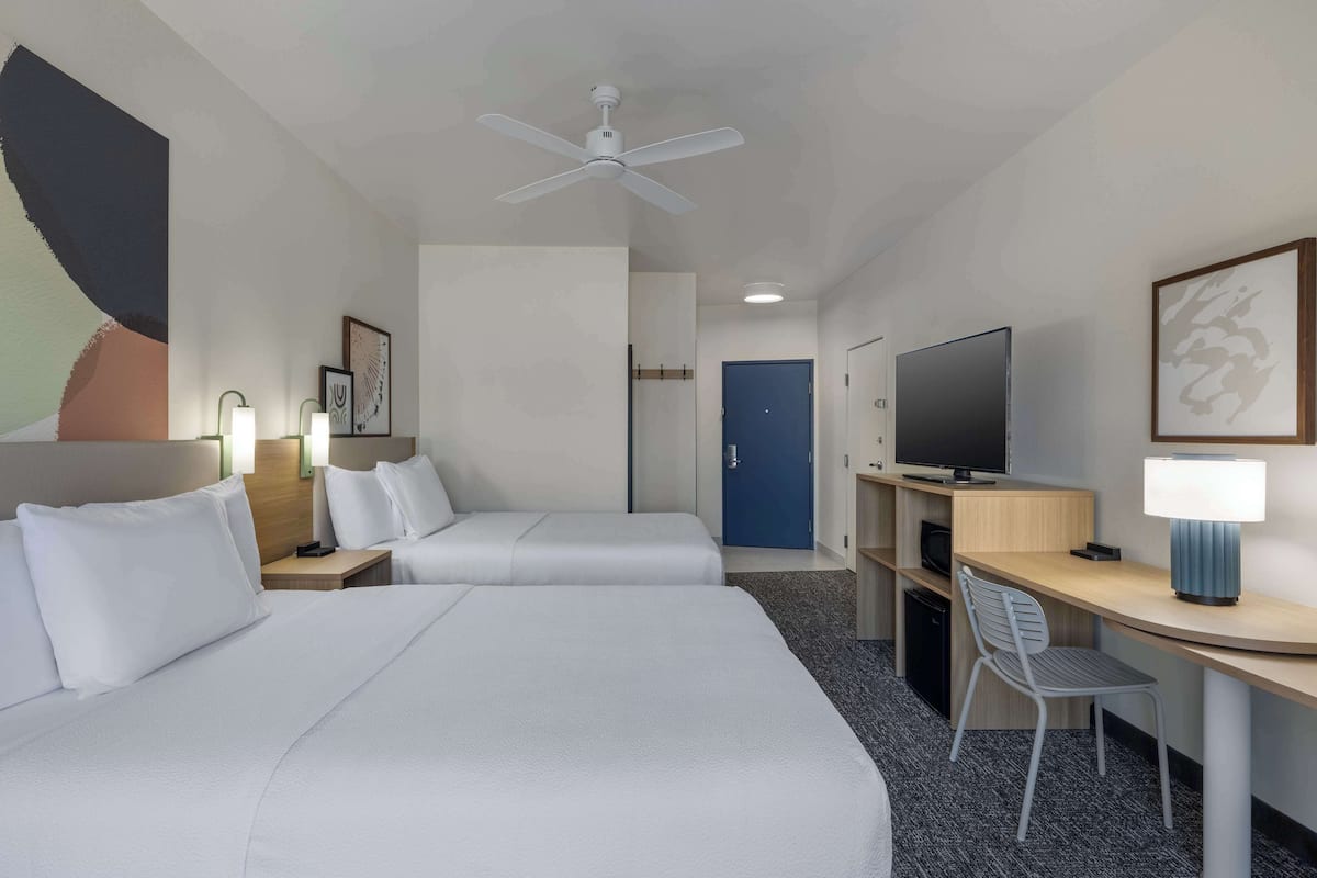 A hotel room with two beds, a desk, a chair, a television, and a ceiling fan.