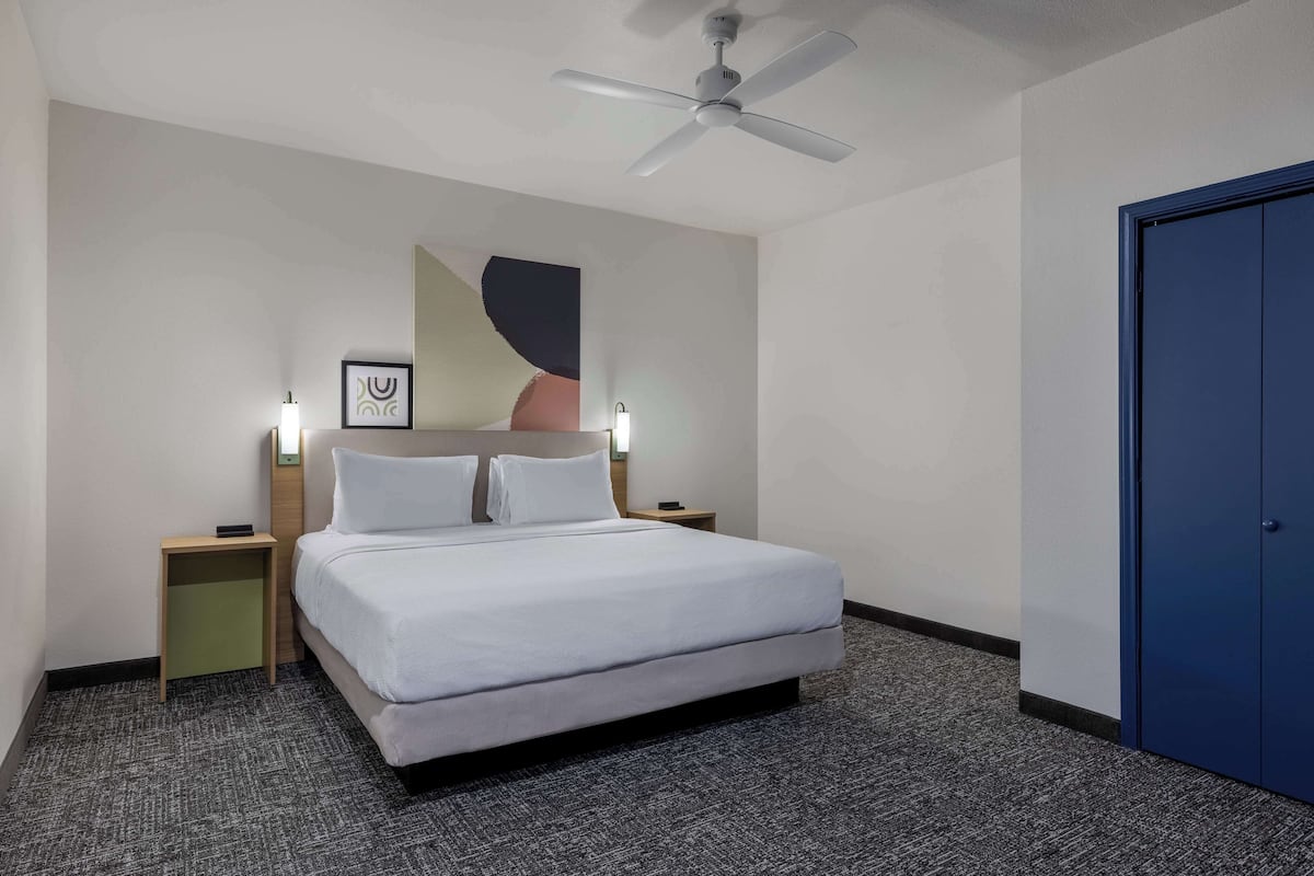 A hotel room with a bed, a ceiling fan, a nightstand, a wall-mounted light, and a door.
