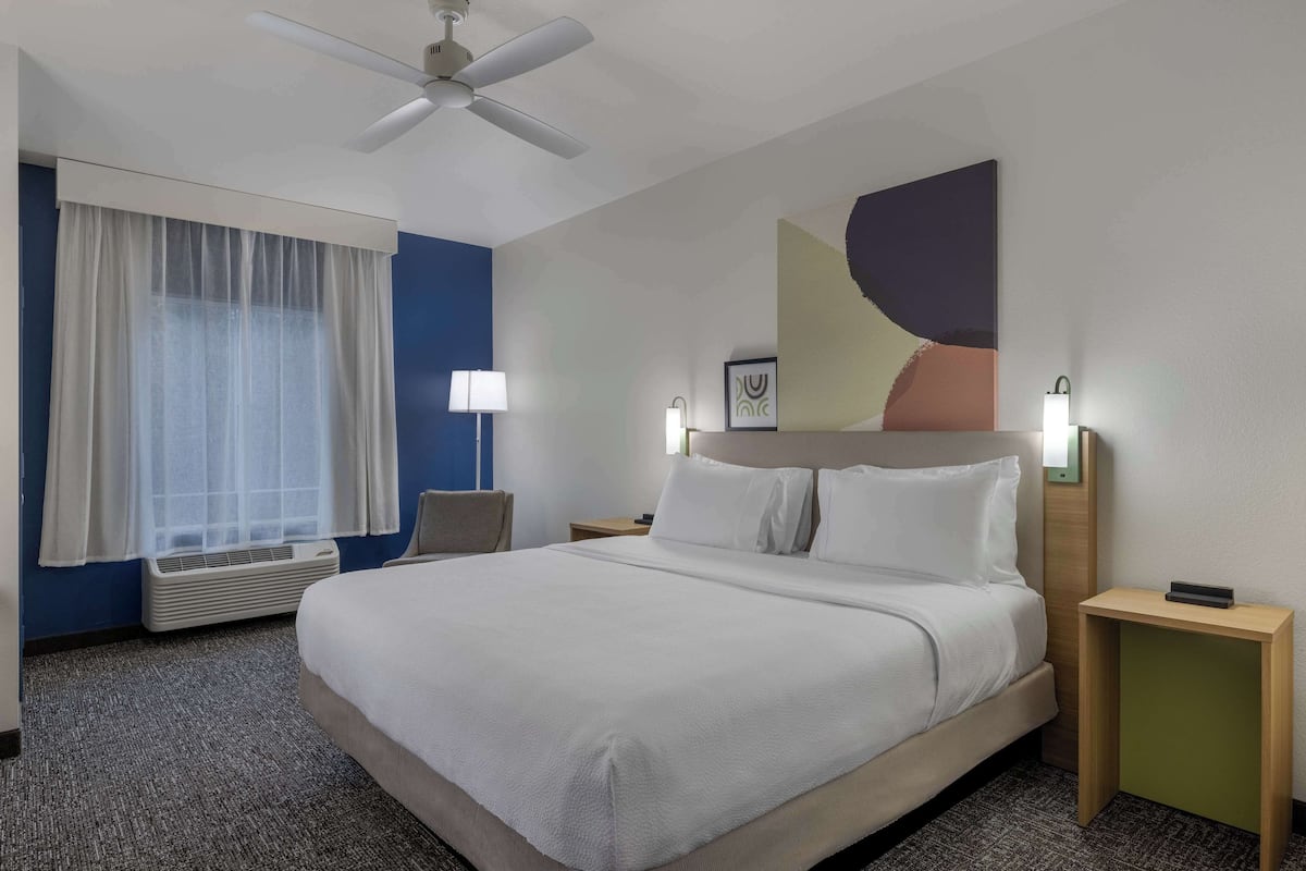 Una habitación de hotel con cama, ventilador de techo, ventana con cortinas, mesita de noche con lámpara y un cuadro en la pared.