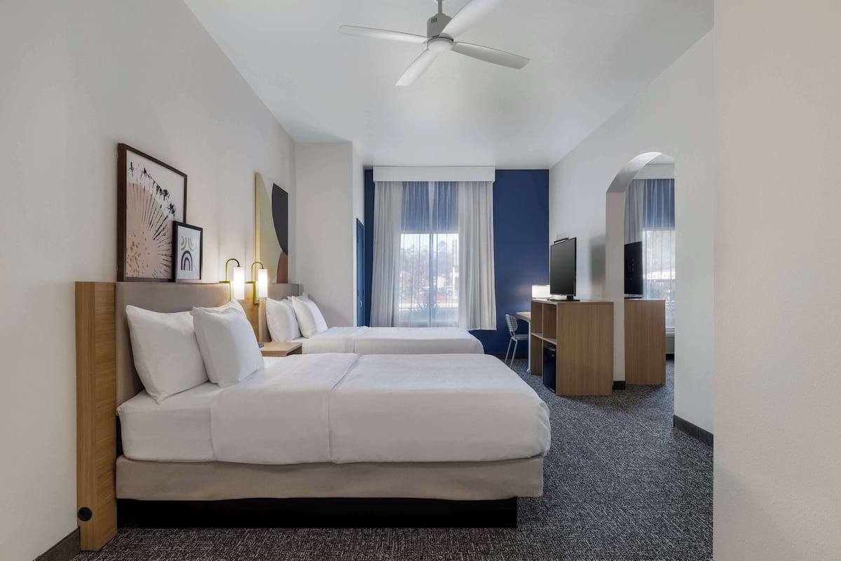 A hotel room with two beds, a ceiling fan, a desk with a chair, and a television.