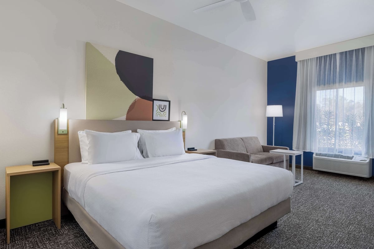Habitación de hotel con una cama grande, un escritorio, una silla, una obra de arte en la pared, una ventana con cortinas y un equipo de aire acondicionado.