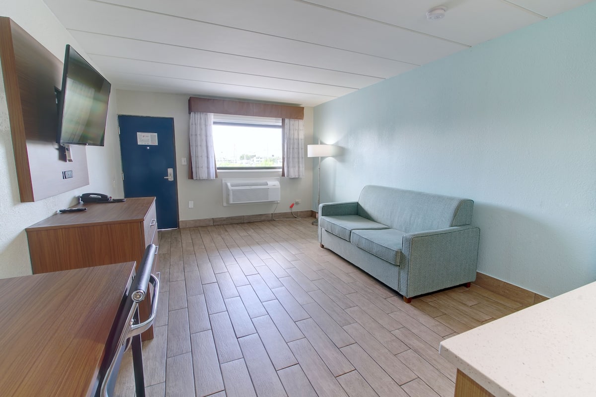 A room with a wooden desk, a television mounted on the wall, a blue door, a window with curtains, a light-colored sofa, and a floor heater.