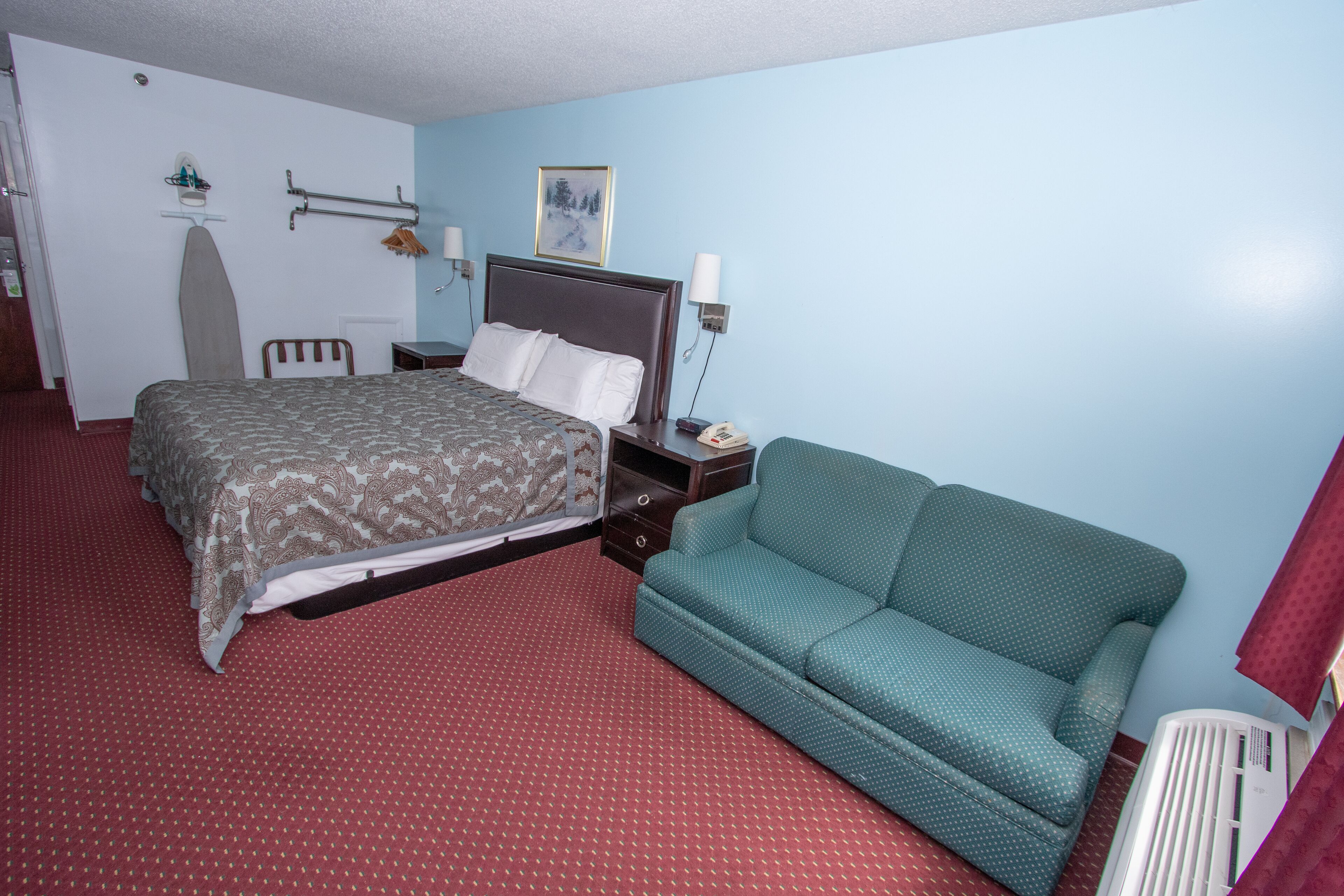 A hotel room with a bed, a green sofa, a red carpet, a wall-mounted heater, and a painting.