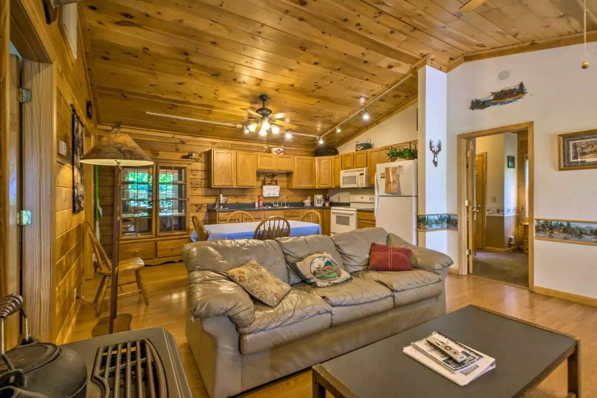 A cozy cabin interior with a wooden ceiling, a leather sofa, a dining table, and a kitchen area.
