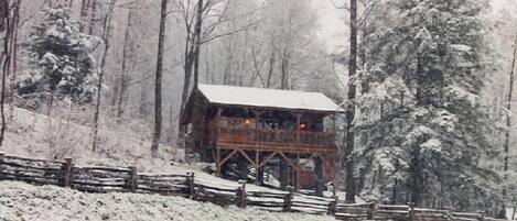 A wooden cabin with a balcony, surrounded by snow-covered trees and a wooden fence.