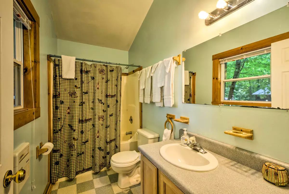 Un baño con cortina de ducha a cuadros, un inodoro, un lavabo con espejo y una ventana con vistas a árboles.