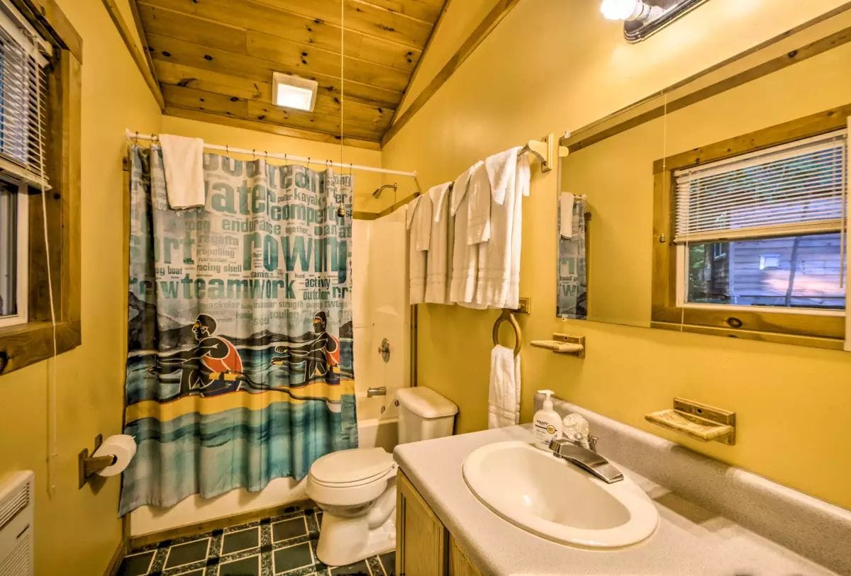 A bathroom with a wooden ceiling, a shower curtain with a nautical theme, a toilet, a sink, and a window with blinds.