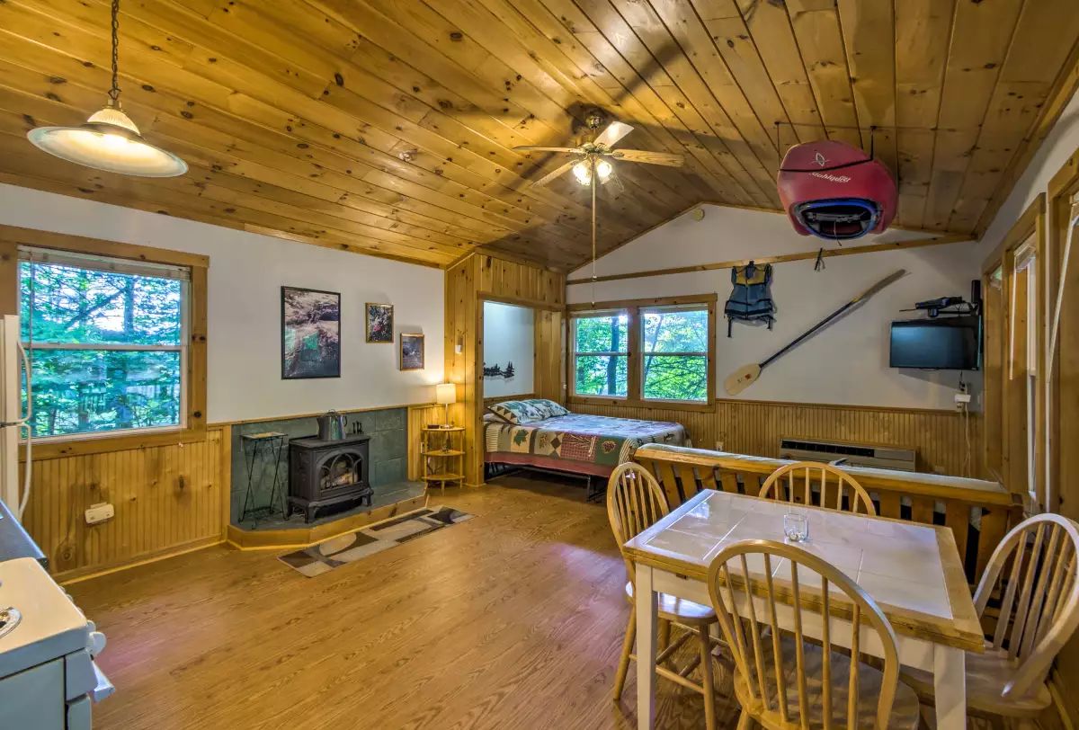 A cozy cabin with a wooden interior, a fireplace, a dining area, and large windows offering views of the forest.