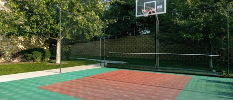 An outdoor basketball court with a green and red playing surface, surrounded by a fence and trees.