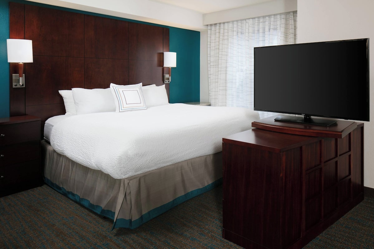 A hotel room with a large bed, a flat-screen TV, a wooden headboard, and a nightstand with a lamp.