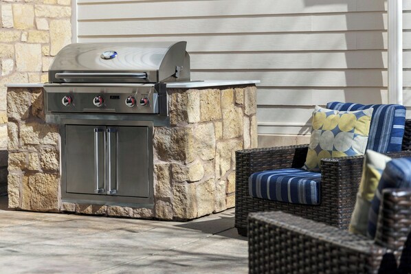 An outdoor grill with a stone base, a wicker chair with cushions, and a stone wall in the background.