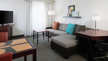 A hotel room with a sofa, ottoman, TV, table, chairs, and a lamp.