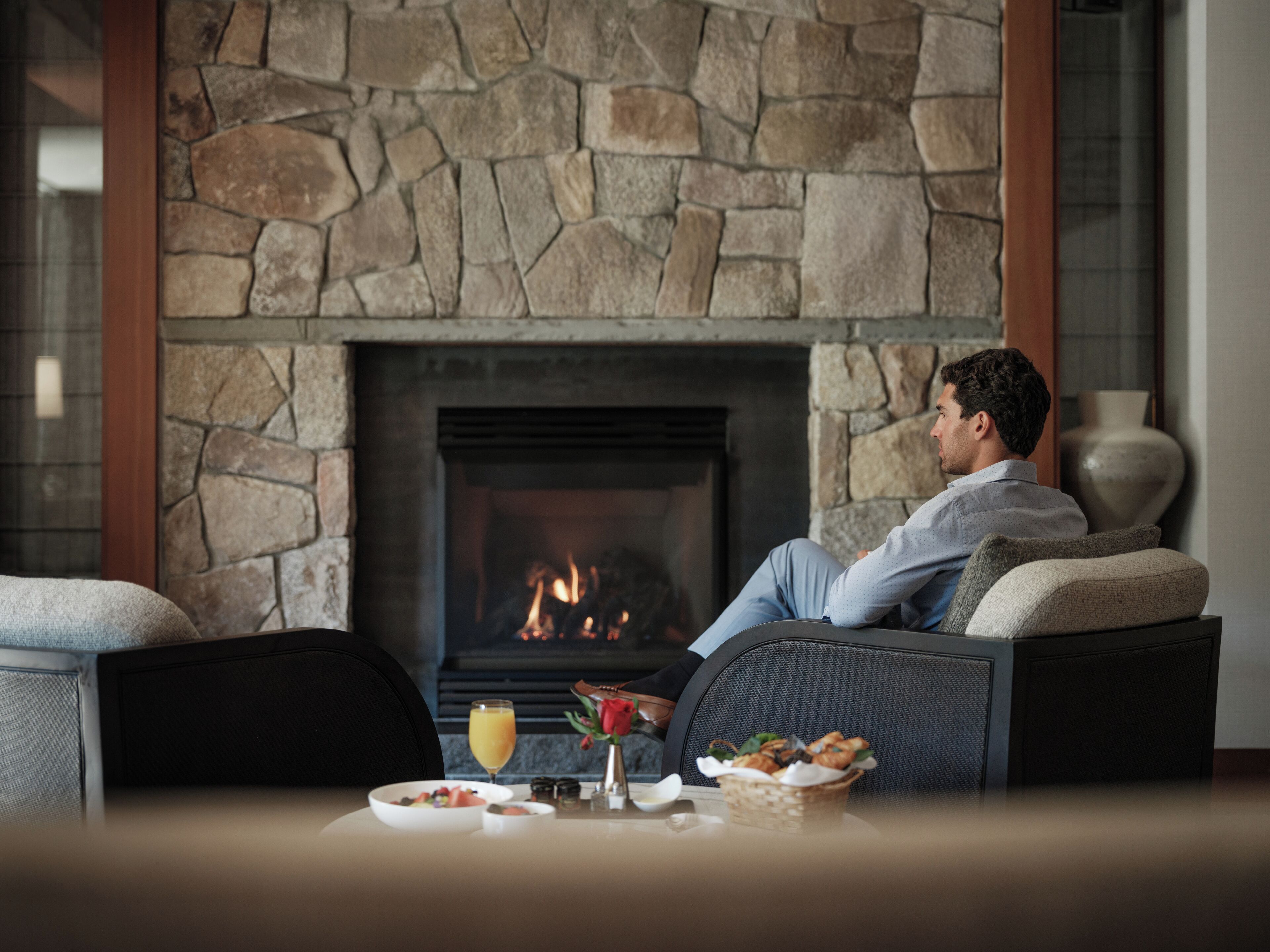 A man relaxing on a sofa by a stone fireplace with a glass of orange juice and a basket of food on the table.