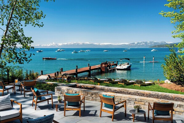 Een terras aan het water met buiten zitplaatsen en uitzicht op boten op een meer, met bergen op de achtergrond.