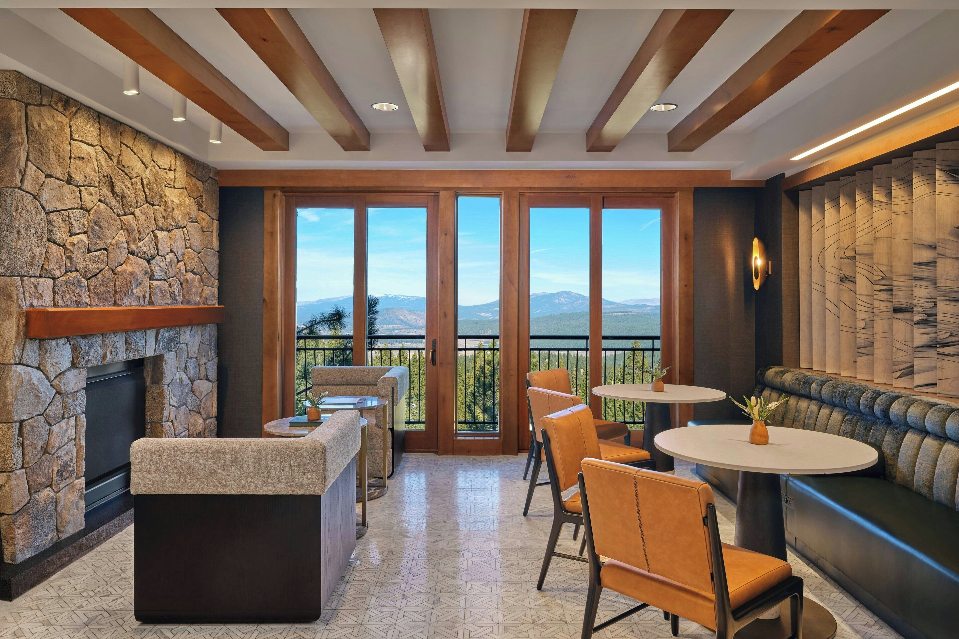 A spacious indoor area with a stone wall, a fireplace, a seating area with a sofa and chairs, and large windows offering a view of mountains.