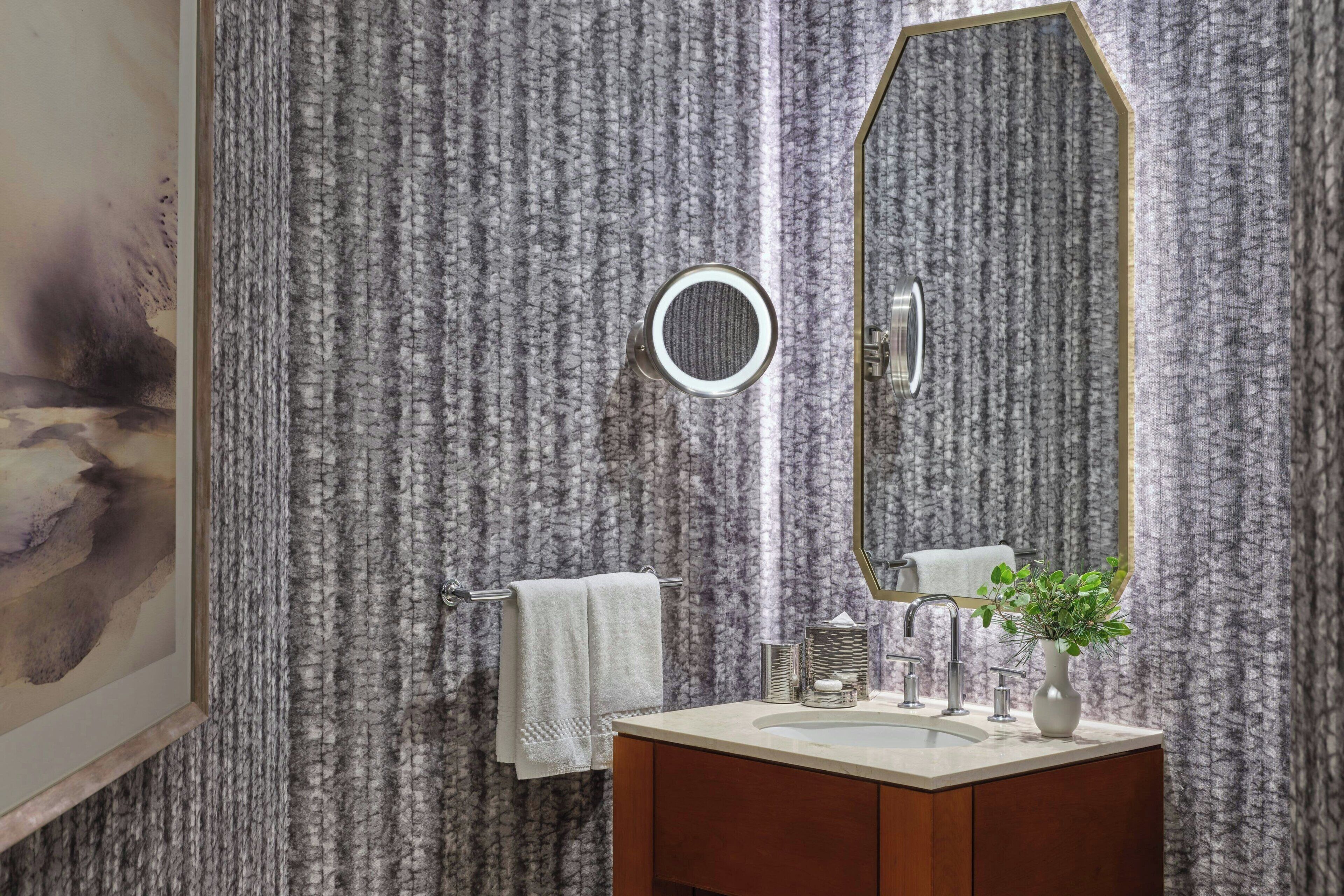 Un baño con pared de diseño de mármol, un espejo redondo, un mueble de tocador de madera y una toalla blanca.