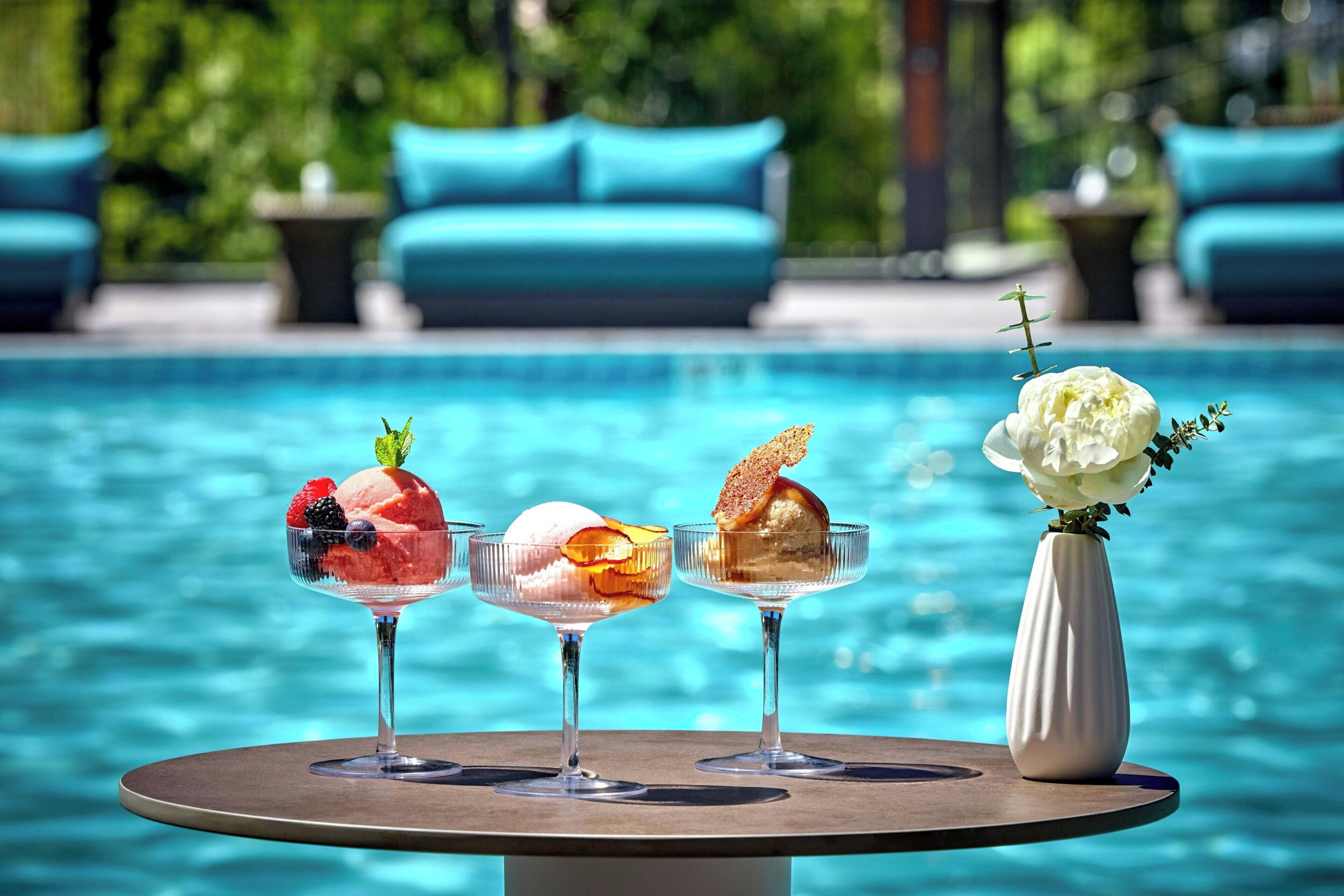 A poolside table with three dessert glasses, a vase with white flowers, and a pool in the background.