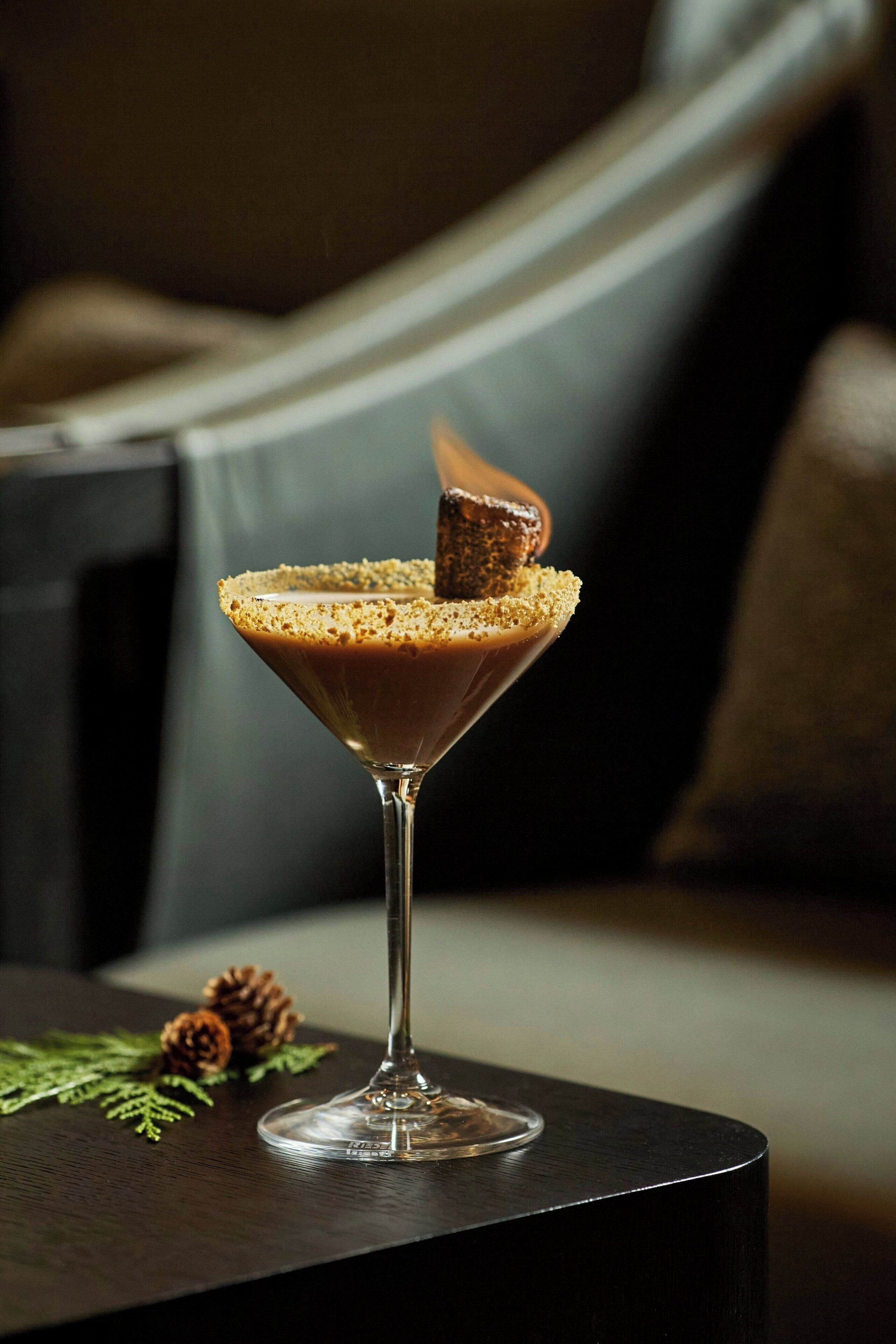 A martini glass with a frothy top and a piece of bread on the rim, placed on a dark table with a pine cone and greenery beside it.