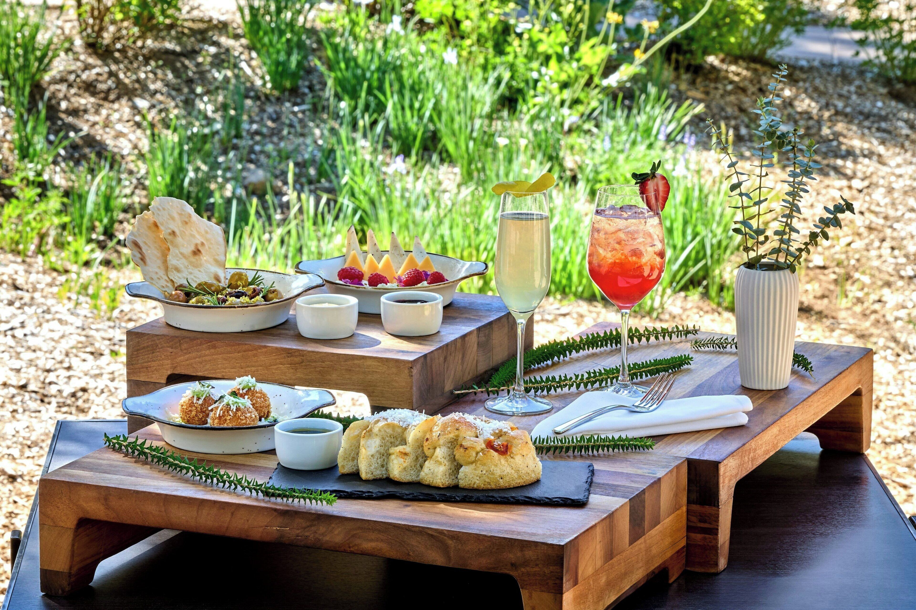 Mobiliario de comedor al aire libre con diversos platos, bebidas y una planta decorativa.