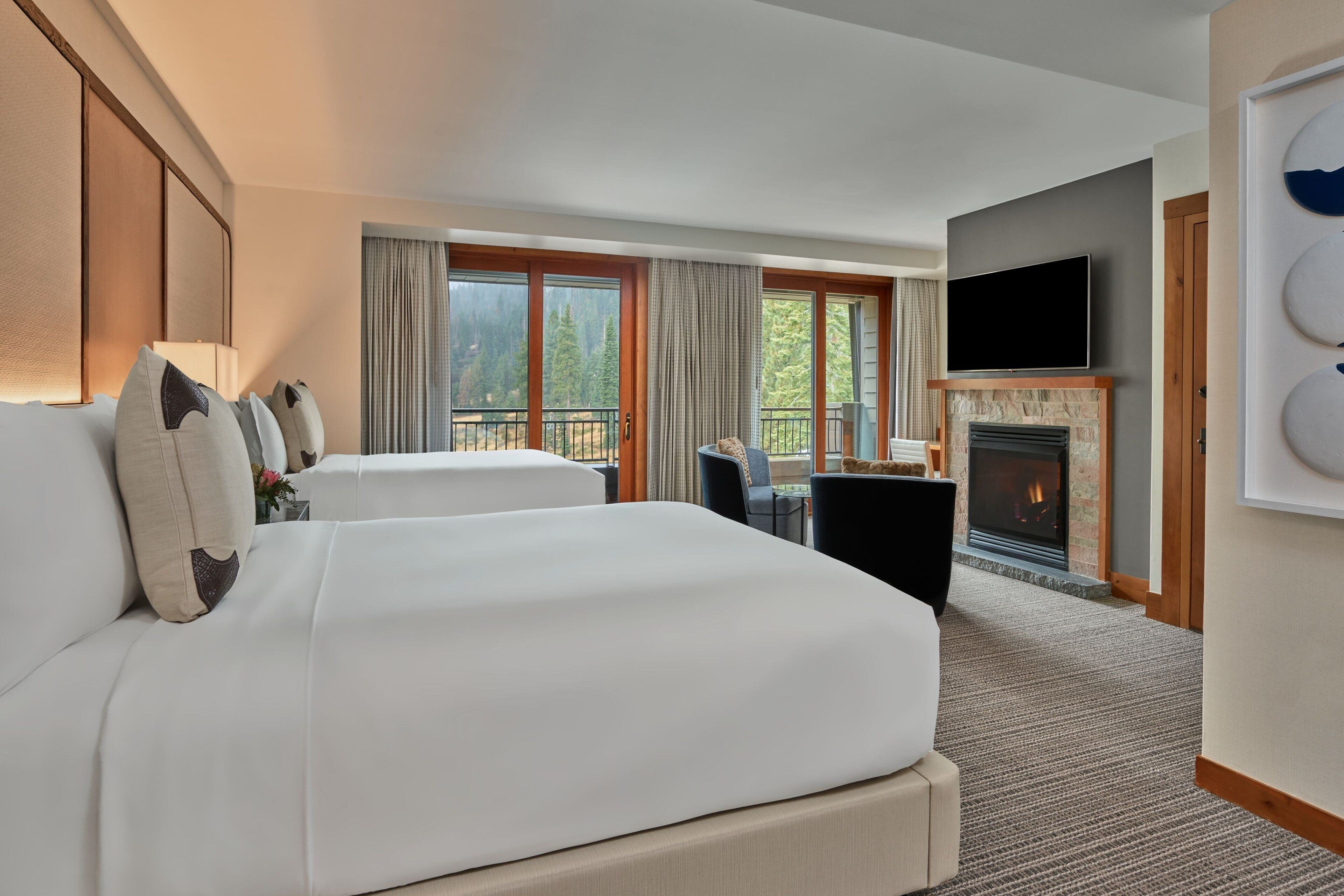 Habitación de hotel con una cama grande, chimenea, televisor y balcón con vistas a árboles.