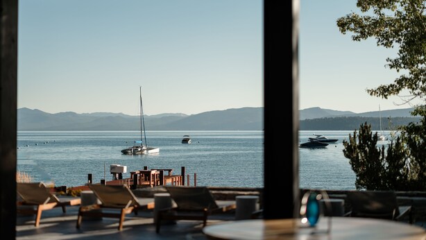 A view from inside a building looking out to a body of water with boats, a sailboat, and a dock with chairs and tables. - The Ritz-Carlton, Lake Tahoe (Truckee)