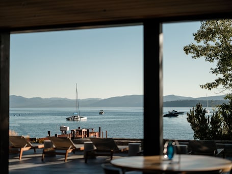 A view from inside a building looking out to a body of water with boats, a sailboat, and a dock with chairs and tables.