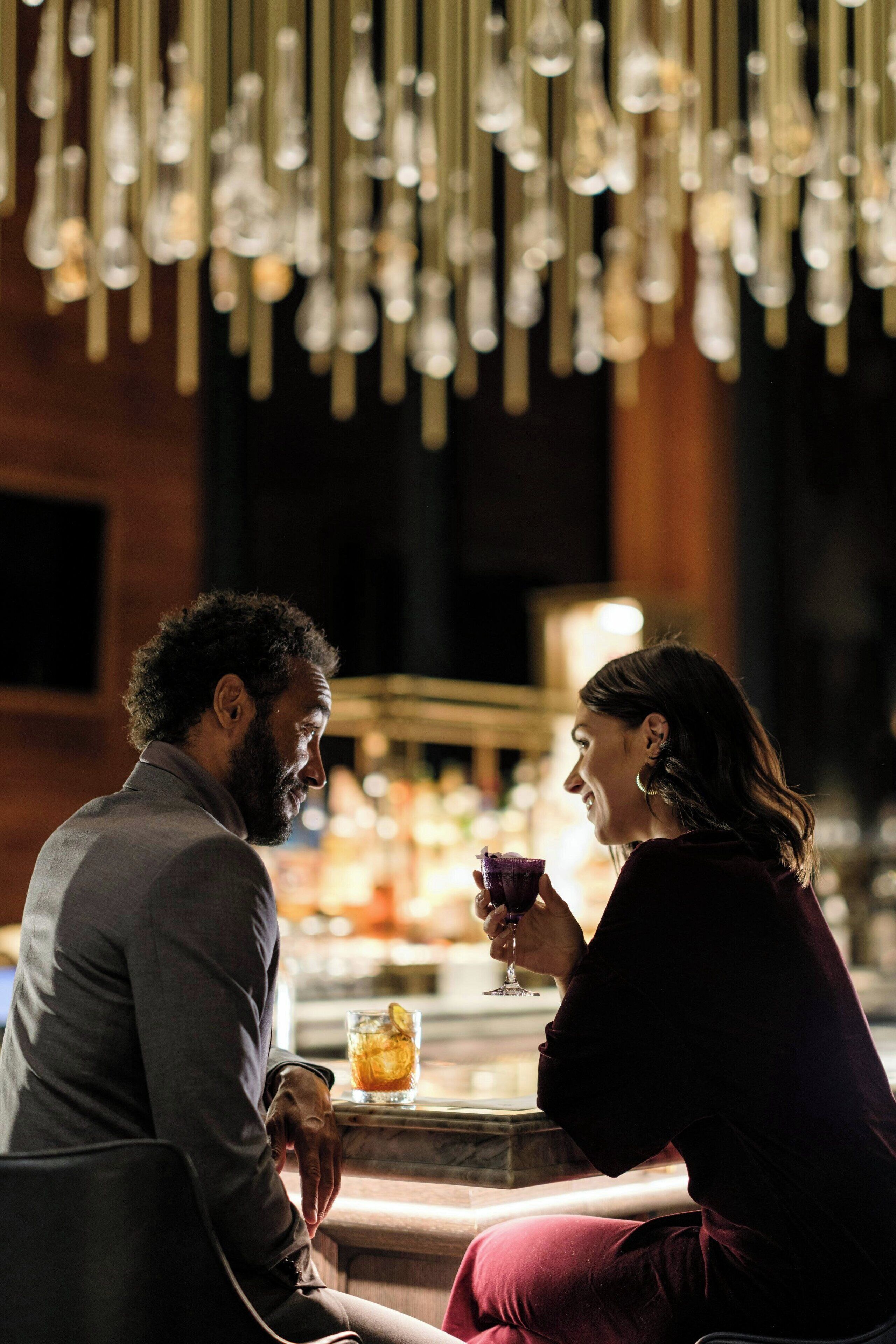 Um casal sentado em um bar, cada um com uma bebida na mão, uma garrafa de uísque e um copo de água no balcão.