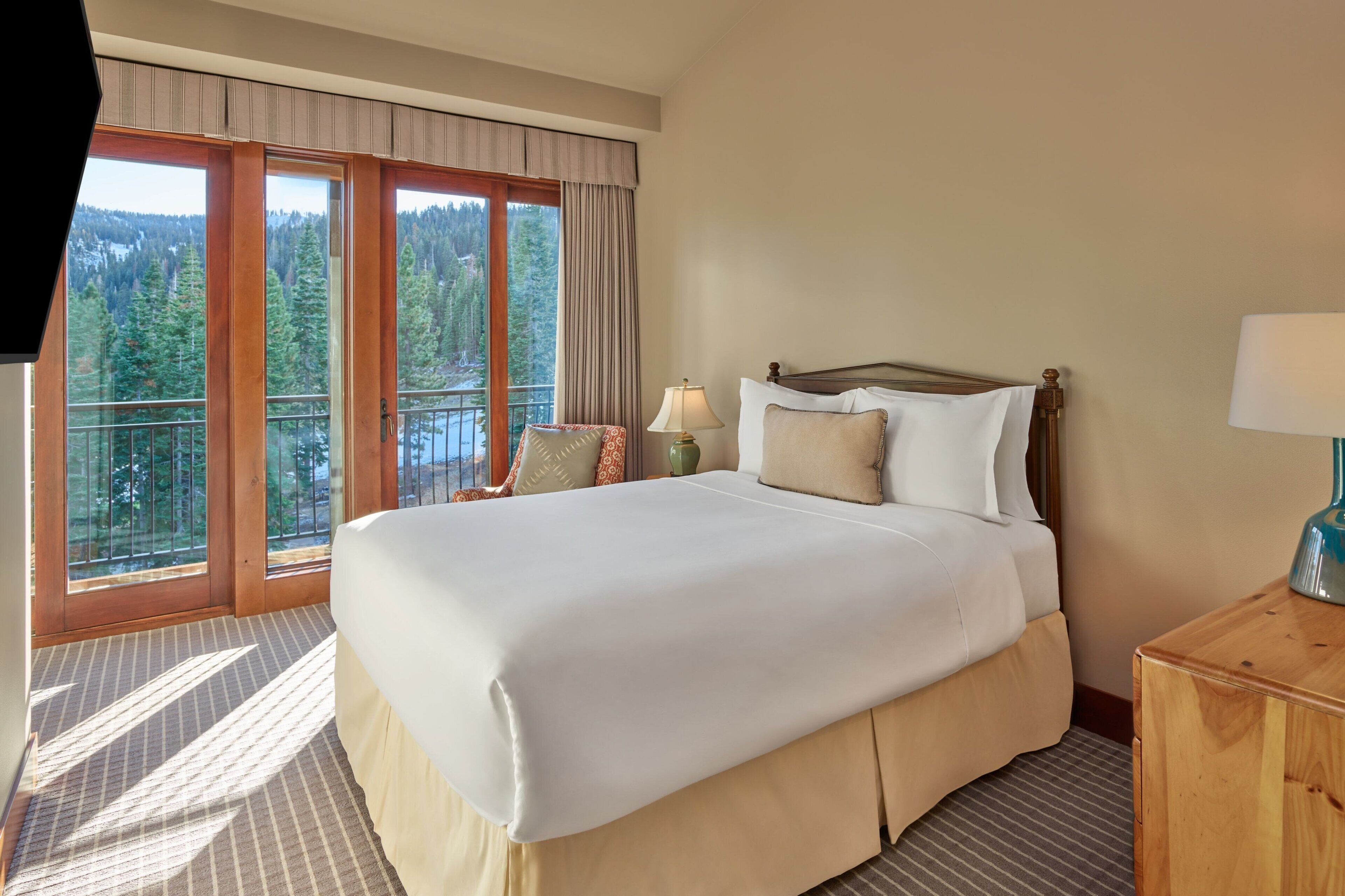 Ein Hotelzimmer mit einem großen Bett, einem Nachttisch aus Holz, einer Lampe und Blick auf einen Wald durch große Fenster.