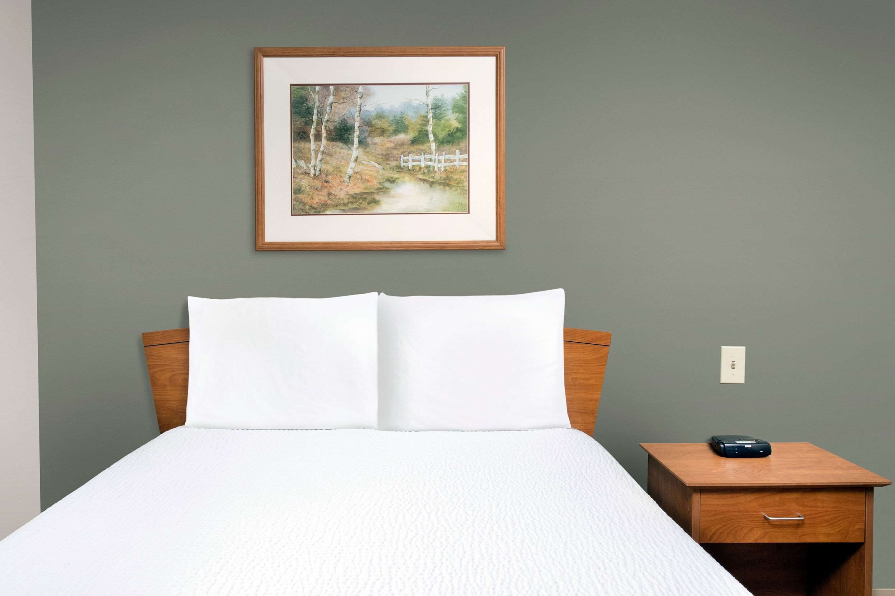 Una cama bien hecha con ropa blanca, un cabecero de madera, una mesita de noche pequeña de madera con un cajón, y un cuadro enmarcado de un paisaje en la pared.