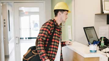 A person in a plaid shirt and yellow hard hat standing at a reception desk with a laptop, tools, and a bag.