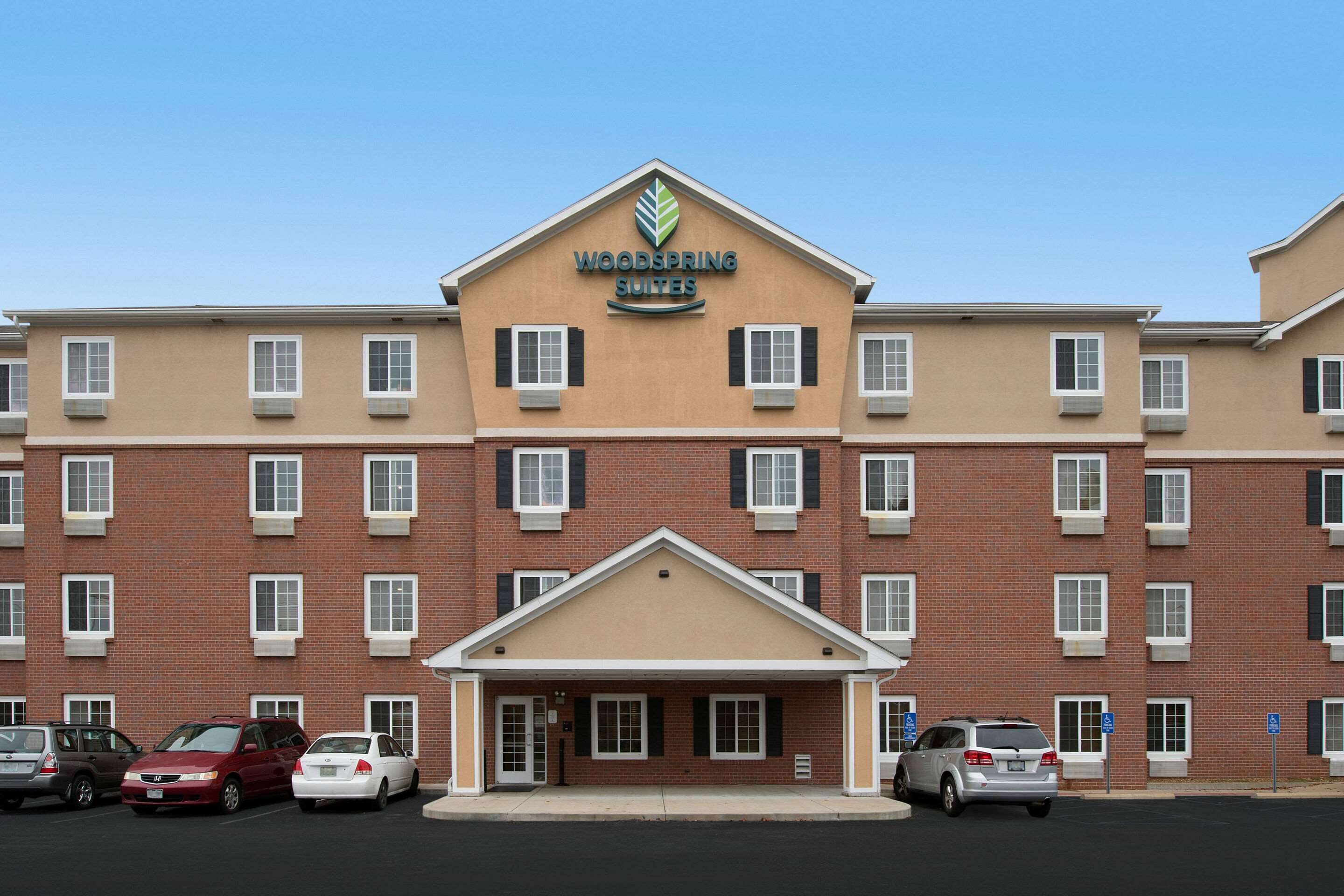 A multi-story hotel with a central entrance, parking lot, and a sign reading "Woodspring Suites".