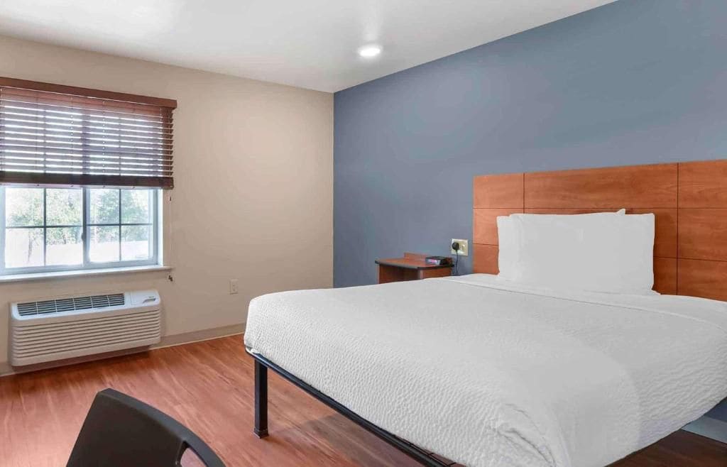 Une chambre d’hôtel avec un lit, une fenêtre avec des stores, une table de chevet et un radiateur mural.