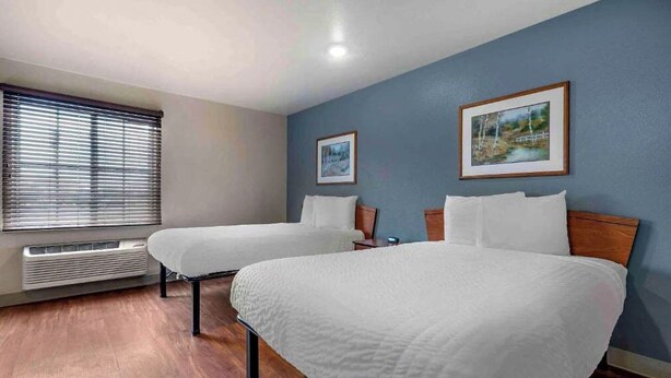 A hotel room with two beds, a window with blinds, a heater, and two framed pictures on the wall. - Studio 6 Extended Stay Bossier City, LA - Shreveport (Bossier City)