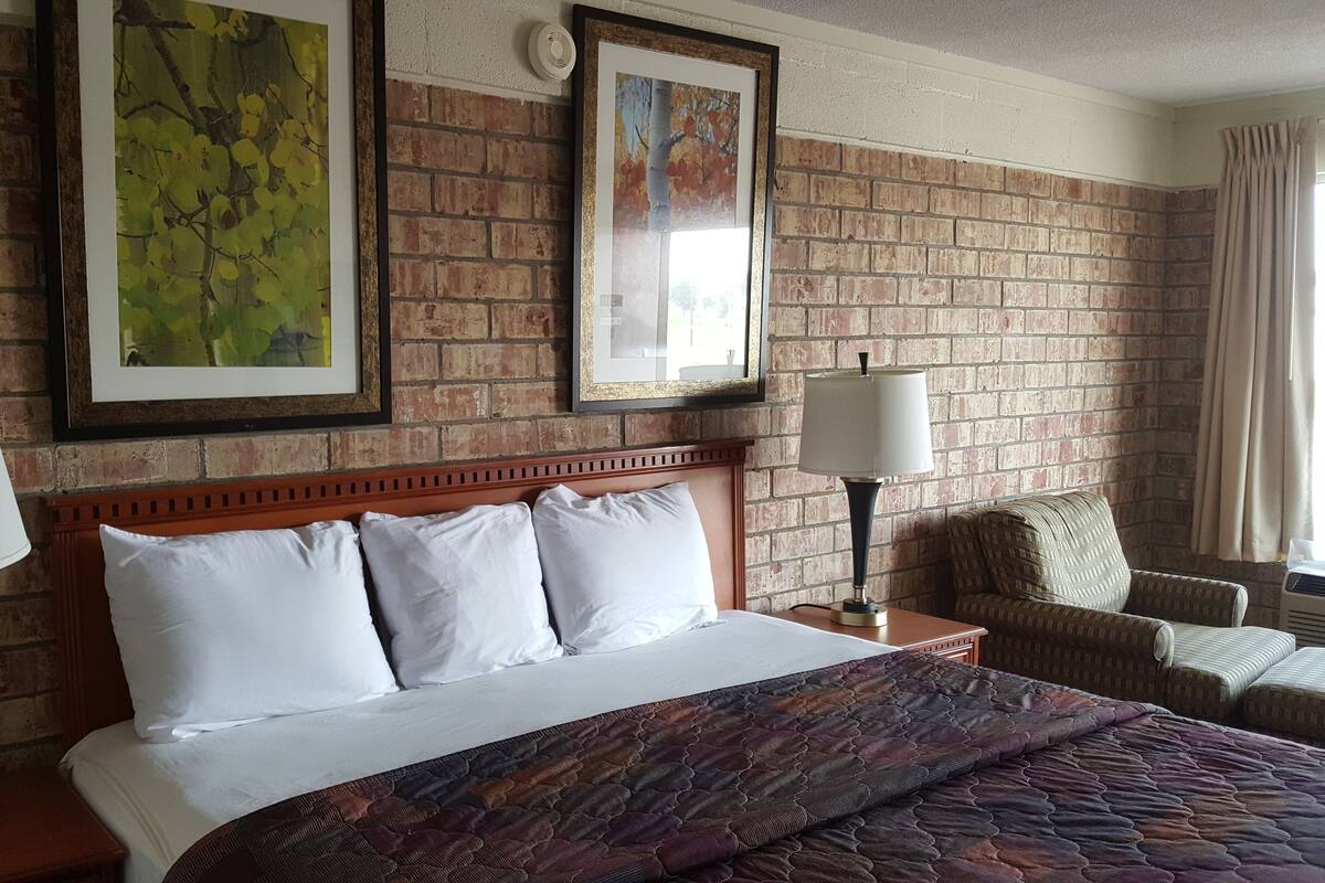 A hotel room with a brick wall, two framed artworks, a bed with white linens, two bedside lamps, a patterned throw blanket, a wooden headboard, and a window with curtains.