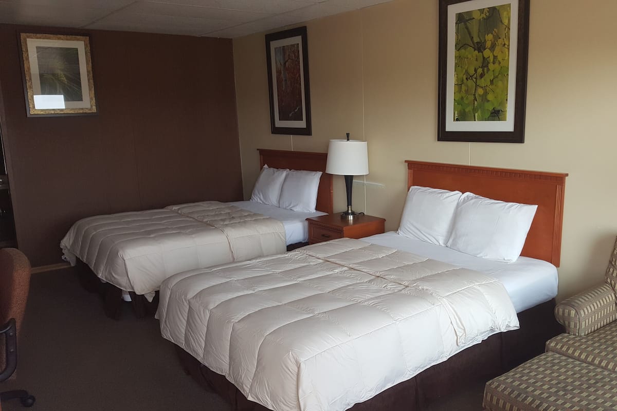 A hotel room with two beds, a nightstand with a lamp, a chair, and framed pictures on the walls.