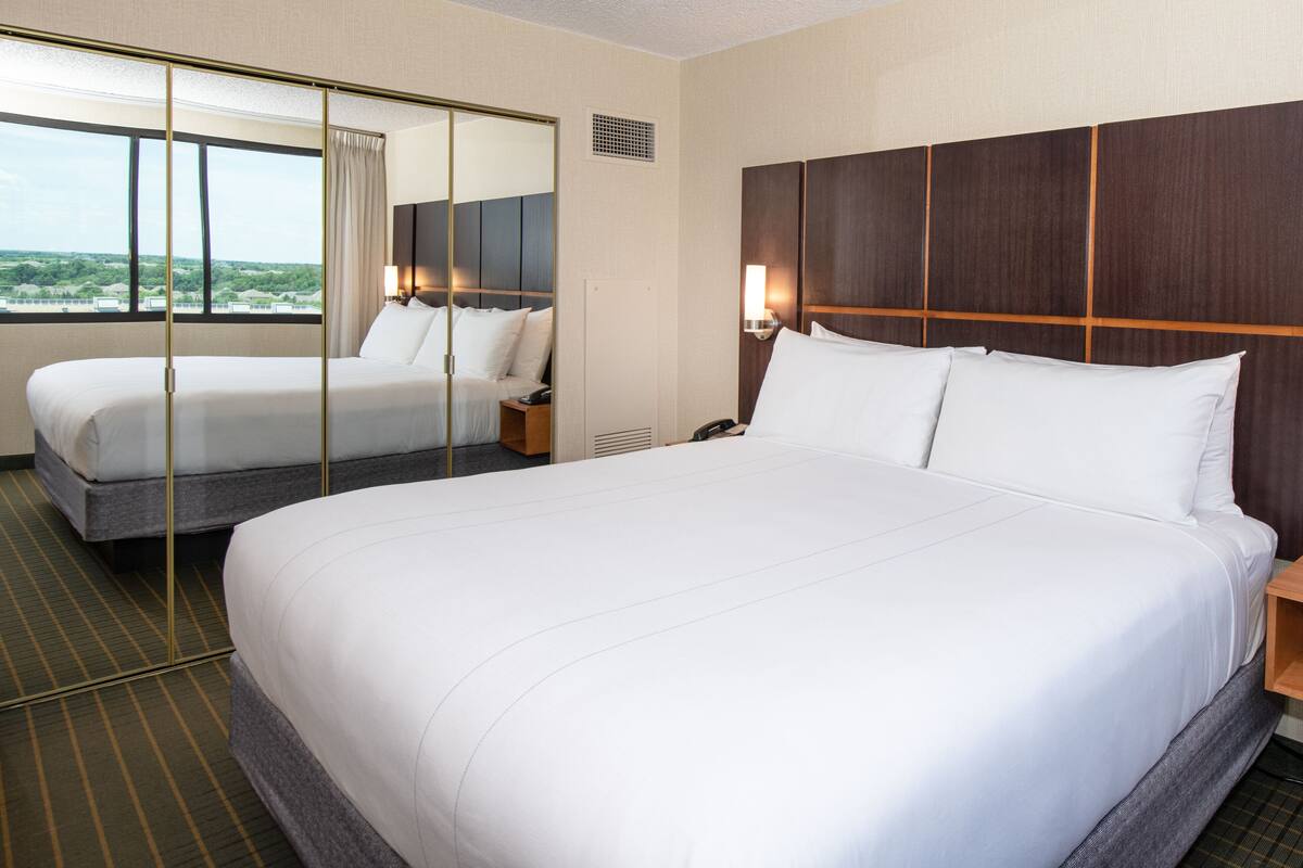A hotel room with a large bed, a bedside table with a lamp, a mirror, and a view of the outdoors.