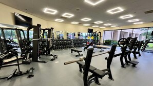 Un gimnasio con diversos equipos de ejercicio, ventanas amplias y un techo con luces cuadradas.