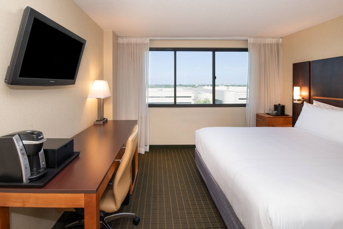 A hotel room with a bed, a desk with a coffee maker, a TV mounted on the wall, and a window with a view of buildings.