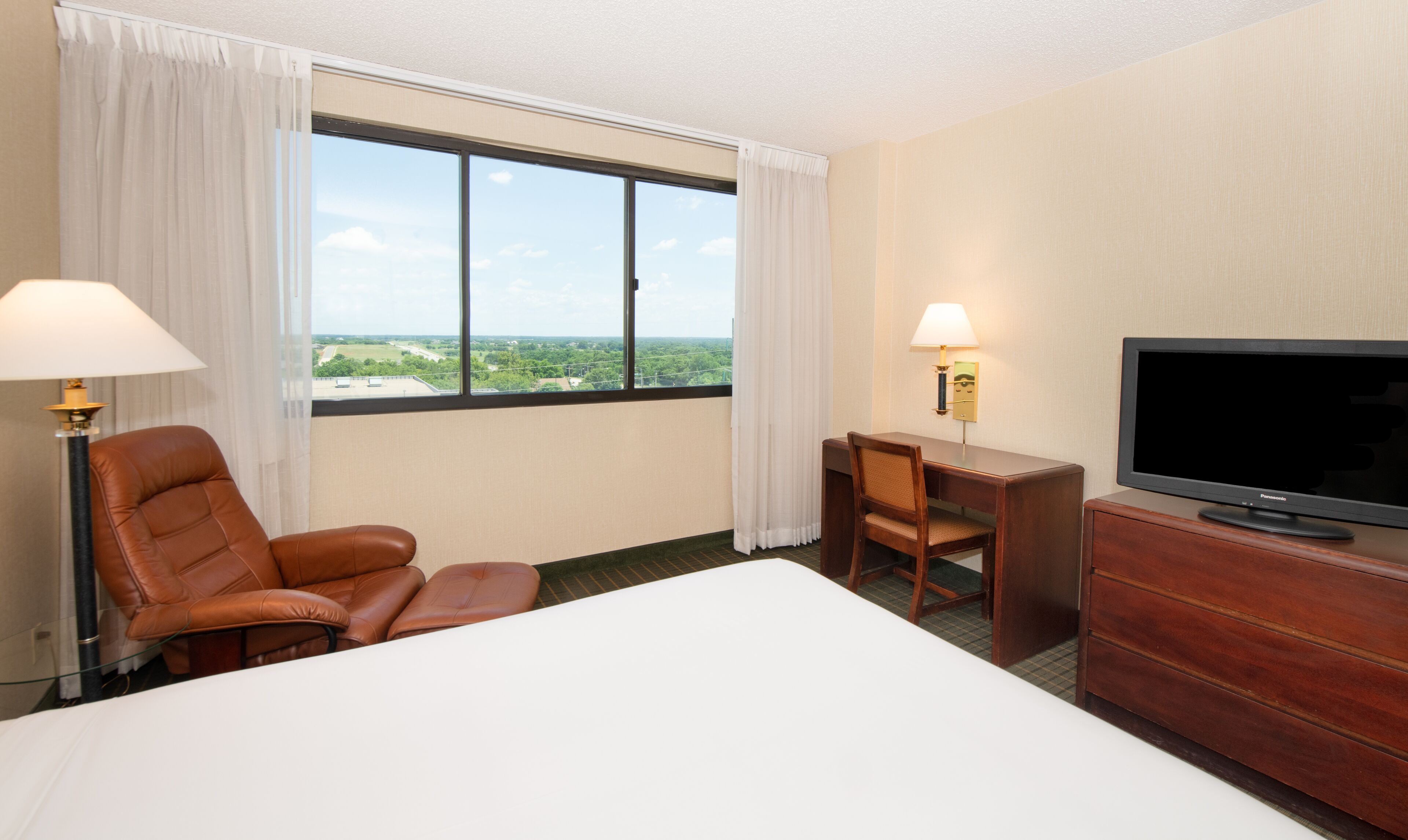 Une chambre d’hôtel avec un grand lit, un bureau, une chaise, une télévision et une fenêtre avec des rideaux.
