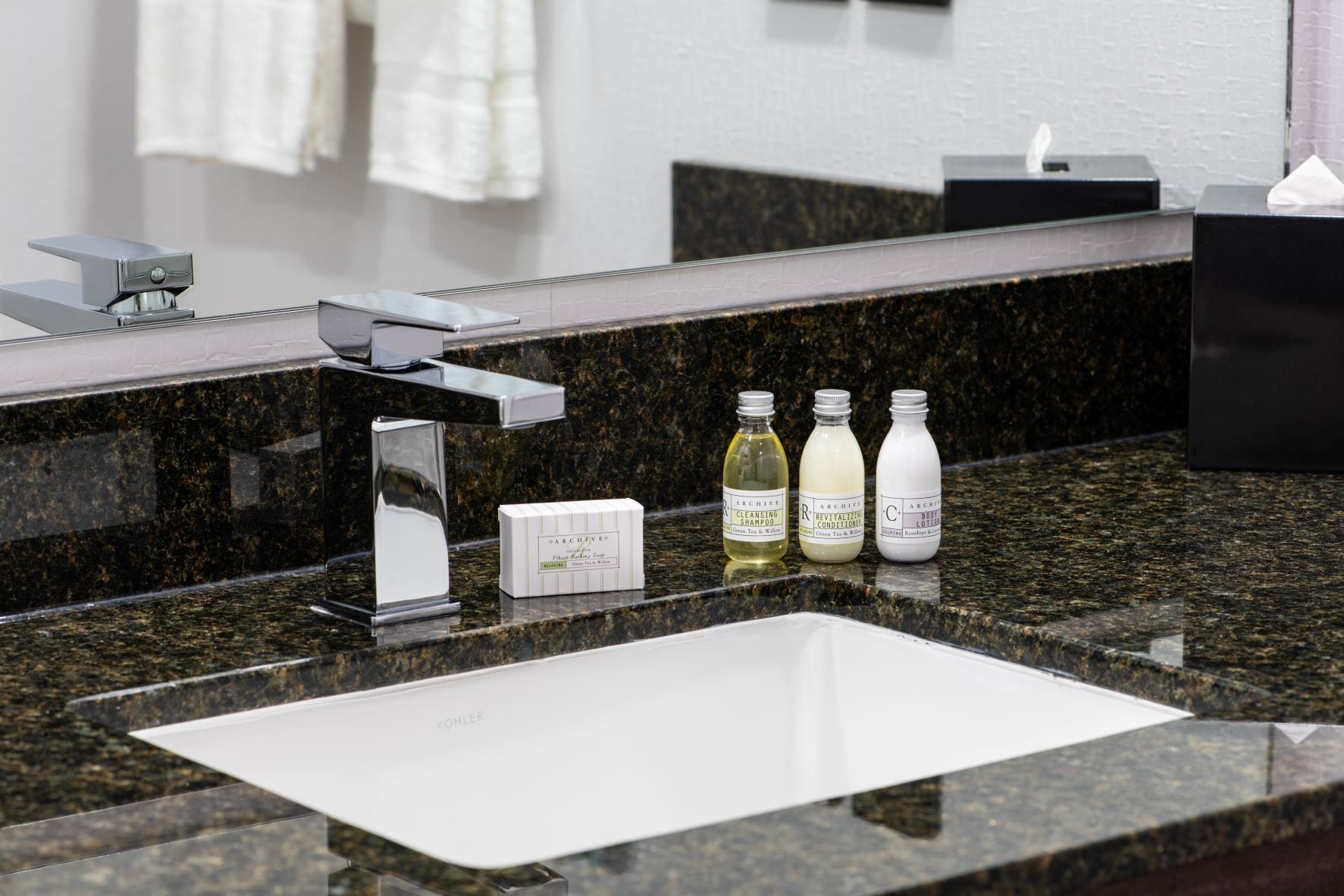 A modern bathroom with a granite countertop, a white sink, and a chrome faucet. There are three bottles of toiletries on the counter.