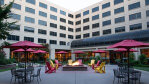 Un patio con mesas al aire libre, una chimenea y un edificio moderno con múltiples ventanas al fondo.