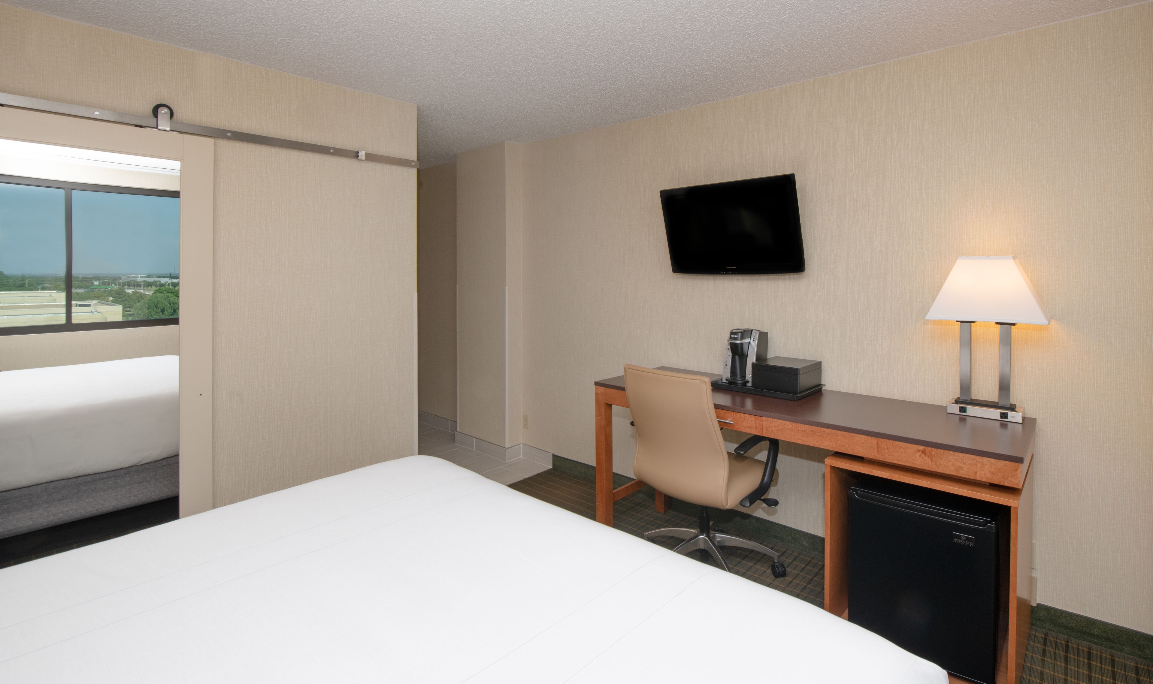 Una habitación de hotel con cama, escritorio con silla, televisor, cafetera y lámpara.