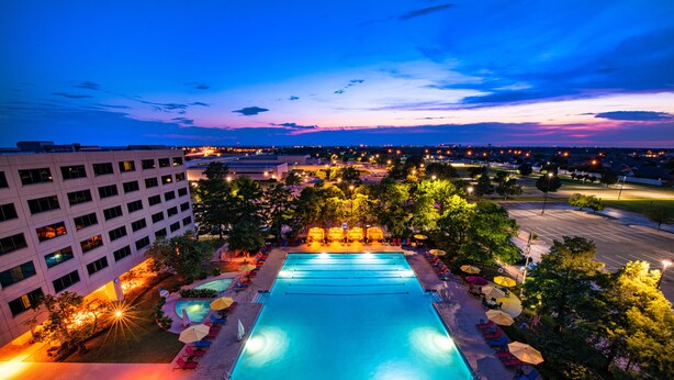 Una gran piscina al aire libre con múltiples carriles, rodeada de sillas de descanso y sombrillas, con un paisaje urbano al atardecer de fondo.