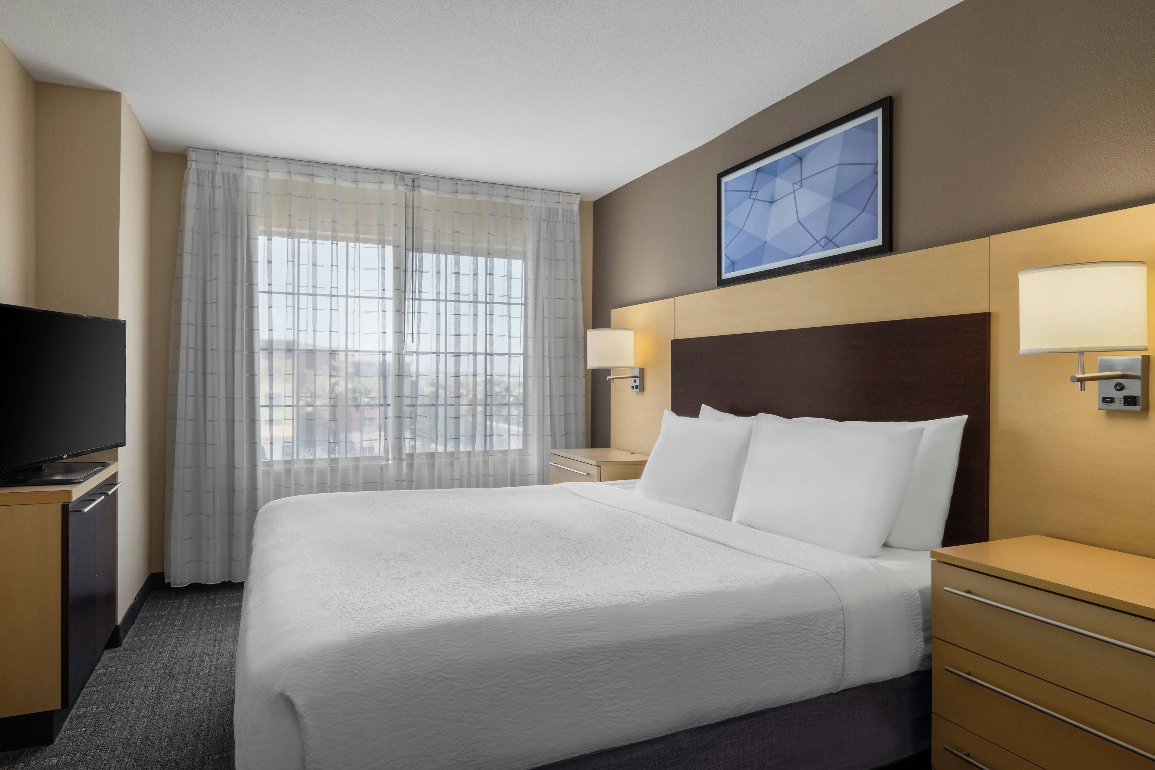 A hotel room with a large bed, a television, a desk, and a window with sheer curtains.