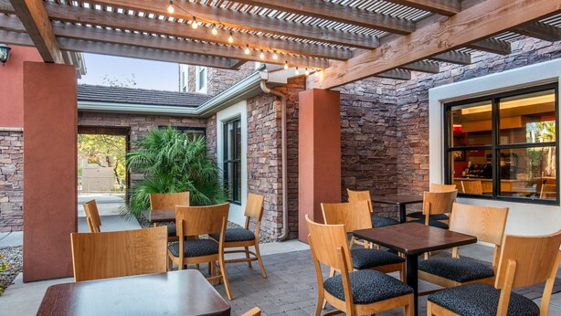 Un patio al aire libre con mesas y sillas de madera, muros de piedra y una pérgola con luces colgantes.